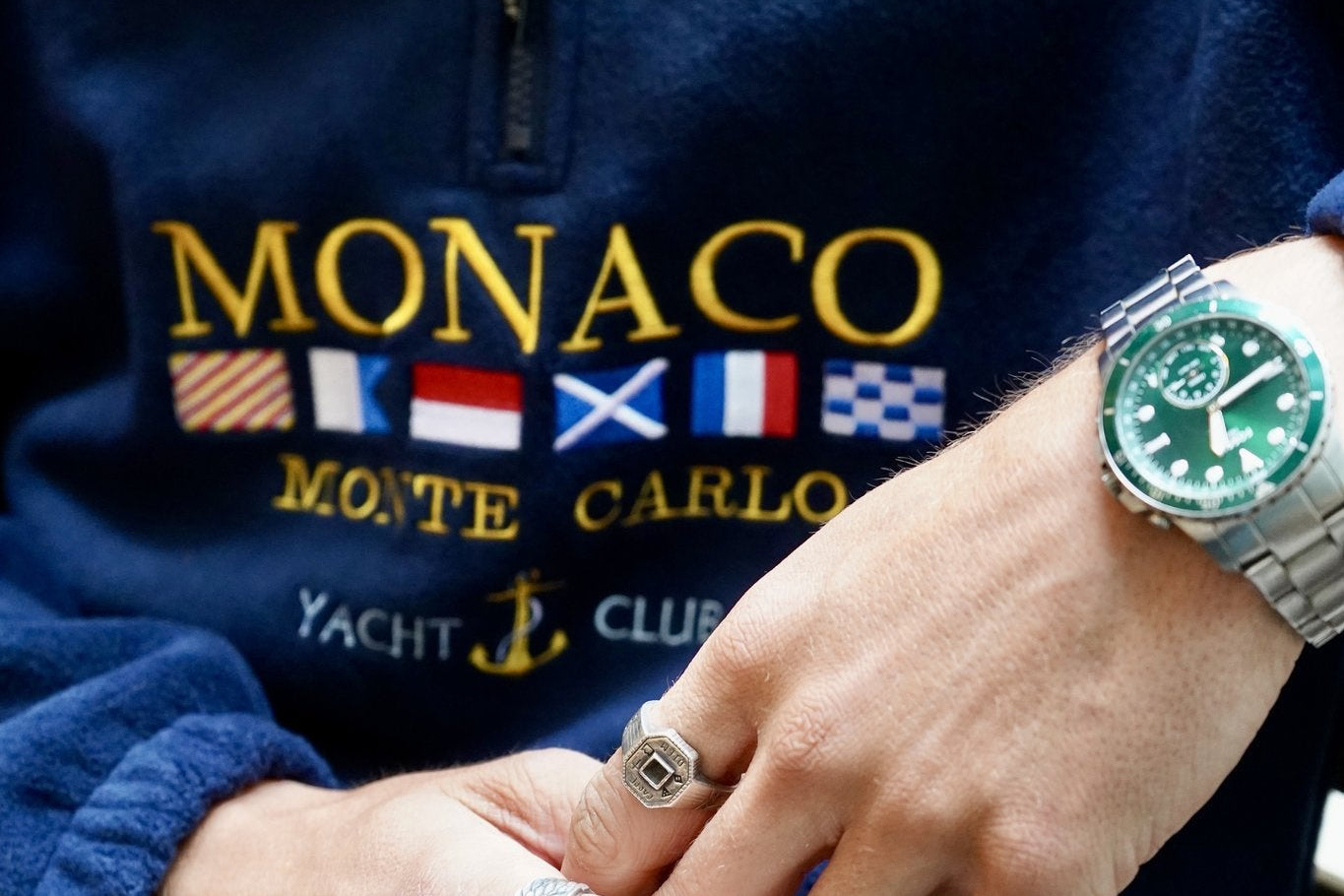 Person wearing a blue Monaco Monte Carlo Yacht Club sweatshirt with a green watch and rings.